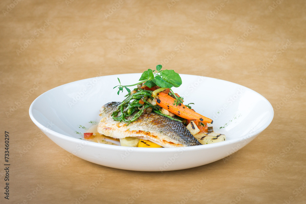 Grilled sea bass fillets with mixed seasonal vegetables and garnish on a white plate with a light brown background Grilled sea bass fillets with mixed seasonal vegetables and garnish on a white plate with a light brown background
