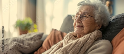 The elderly woman is sitting comfortably on a wooden couch in a cozy room, wearing glasses, a scarf, and a happy smile on her face. photo