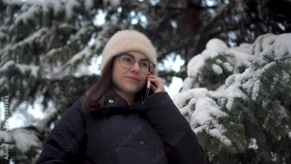 A young woman speaks on the phone near a tall spruce tree in winter. A girl with glasses communicates via cell phone against the backdrop of snowy spruce branches. A young woman speaks on the phone near a tall spruce tree in winter. A girl with glasses communicates via cell phone against the backdrop of snowy spruce branches.