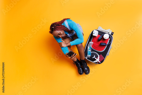 African american girl using mobile phone sitting on the floor in studio, having big trolley bag filled with travelling essentials next to her. Young adult with smartphone preparing to leave.