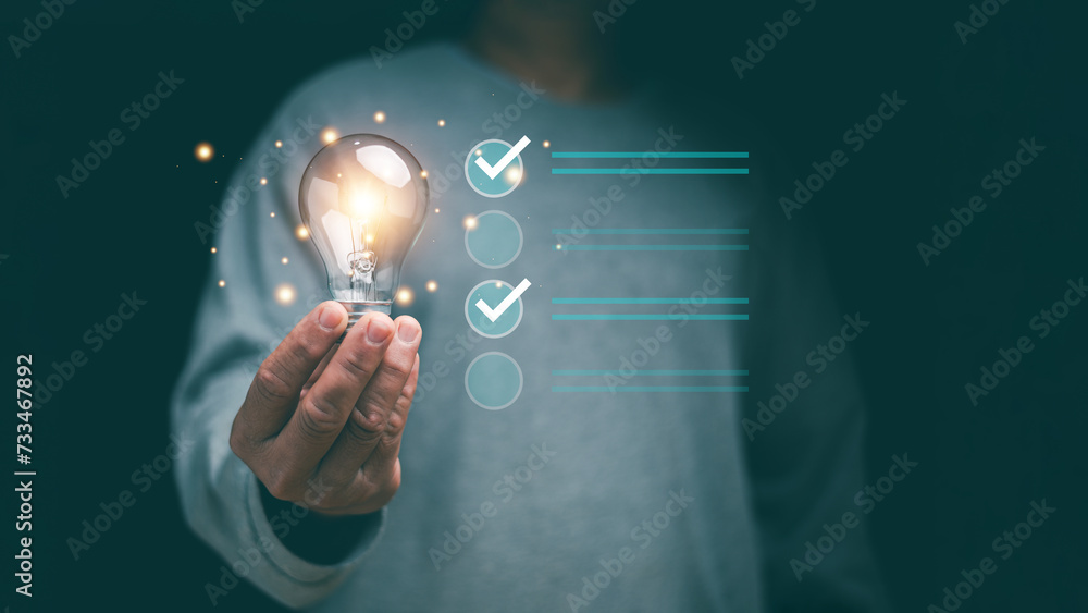 © krungchingpixs - Concept of successful business and thinking solution strategy. Businessman holding glowing light bulb and have checklist for solving problem. Check and solution list to business target.