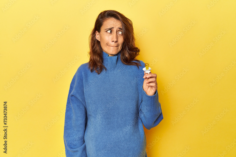 Pregnant woman with pacifier on yellow studio confused, feels doubtful and unsure. Pregnant woman with pacifier on yellow studio confused, feels doubtful and unsure.