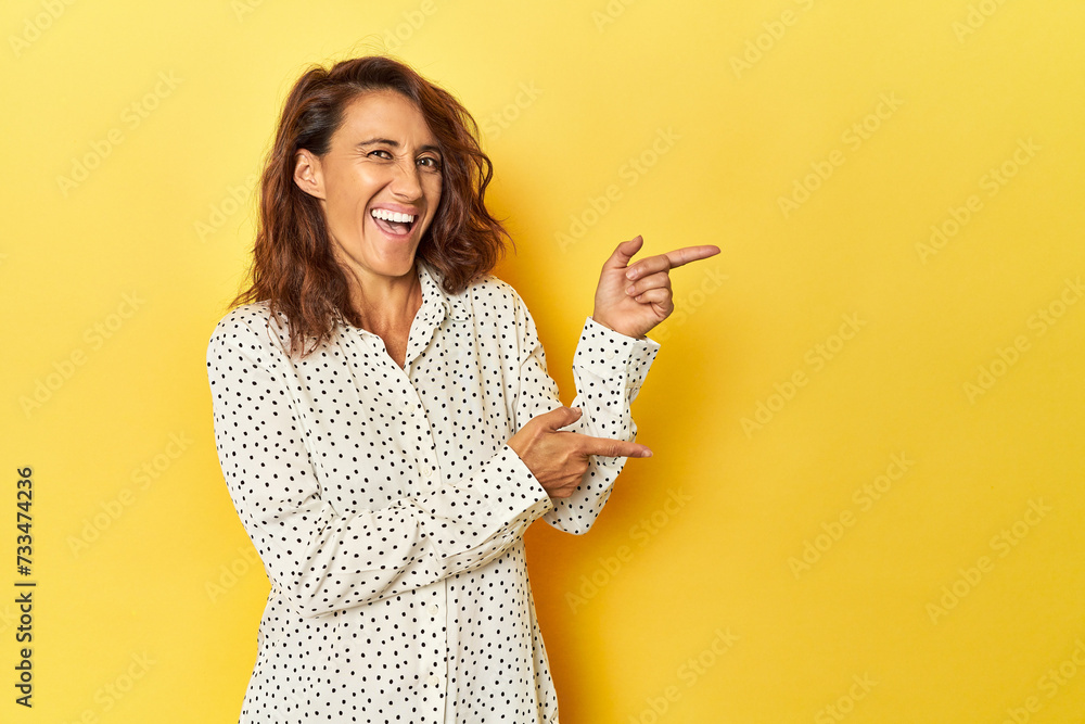 Middle-aged woman on a yellow backdrop pointing with forefingers to a copy space, expressing excitement and desire. - obrazy, fototapety, plakaty 