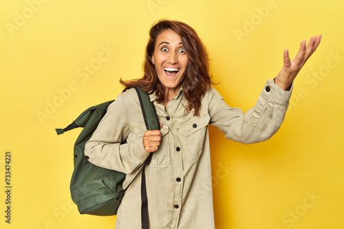 Middle aged woman ready for travel with backpack on yellow receiving a pleasant surprise, excited and raising hands.