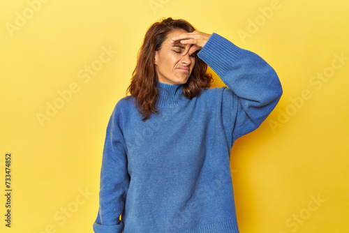 Middle-aged woman on a yellow backdrop having a head ache, touching front of the face. © Asier