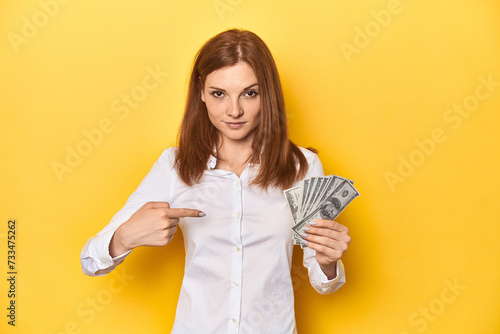 Redhead with dollar bills, cash in hand person pointing by hand to a shirt copy space, proud and confident