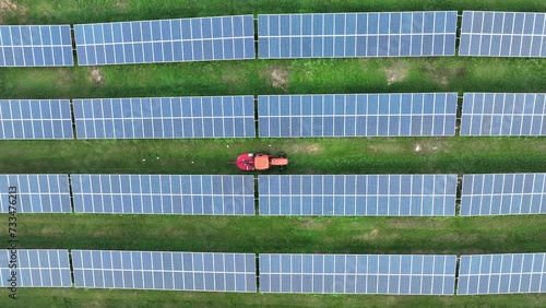 Top down drone view of solar panels in rows with tractor mowing grass between in tropical country. Aerial shot zooming out.