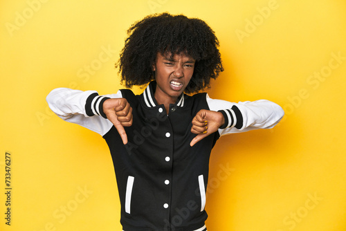 Teen girl in baseball jacket, yellow studio background showing thumb down and expressing dislike.