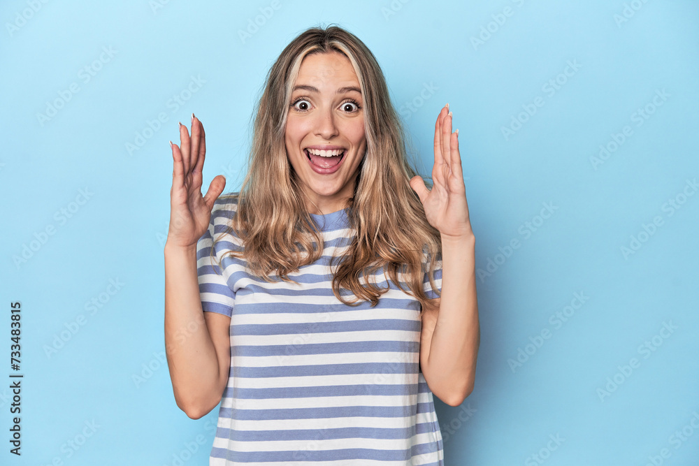Fototapeta premium Blonde young caucasian woman in blue studio celebrating a victory or success, he is surprised and shocked.