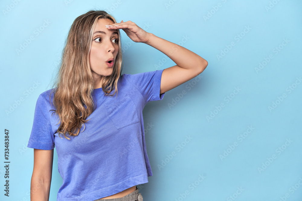 Blonde young caucasian woman in blue studio looking far away keeping hand on forehead. Blonde young caucasian woman in blue studio looking far away keeping hand on forehead.