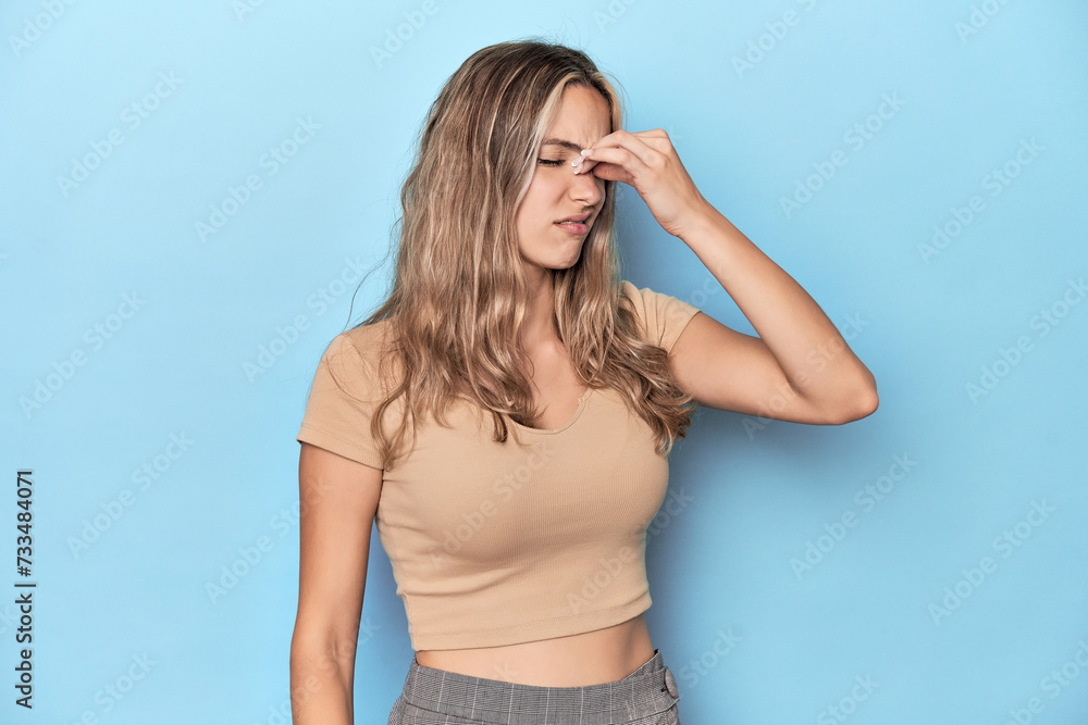 Blonde young caucasian woman in blue studio having a head ache, touching front of the face. Blonde young caucasian woman in blue studio having a head ache, touching front of the face.