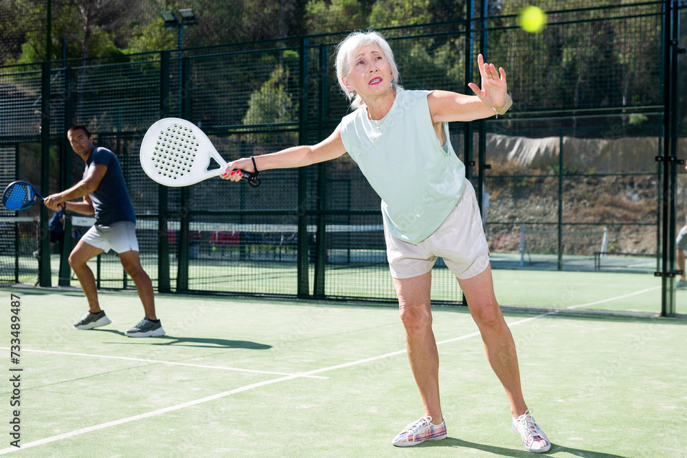 Senior woman in shorts playing padel tennis on court. Racket sport training outdoors.