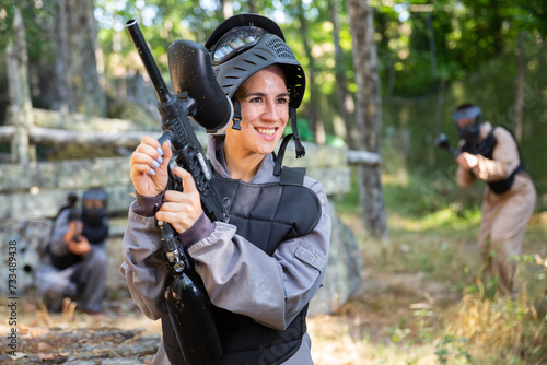 Waist-up portrait of happy latin woman in protective paintball equipment standing on outdoor battleground and smiling.