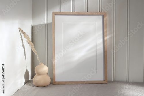 Blank vertical picture frame mock up in sunlight. Modern vase with dry grass festuca. White wooden wall background with floral shadows. Neutral Scandinavian home decor. Nordic interior. photo