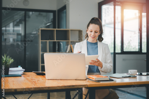 Business analytics concept, Smiling businesswoman using a notebook with a laptop on her desk in a bright, modern office space.