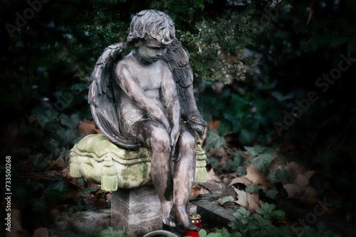 a little angel with wings sits sadly slumped on a stone pillow at a gravestone in a cemetery