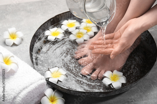 Woman pouring water onto hand while soaking her feet in bowl on light grey floor, closeup. Spa treatment