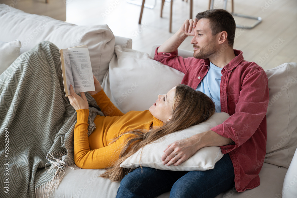 Smiling husband sitting on sofa, beloved wife lying on male knees, reading interesting novel of contemporary author. Loving couple discussing exciting book. Spending weekends, pastime together at home Smiling husband sitting on sofa, beloved wife lying on male knees, reading interesting novel of contemporary author. Loving couple discussing exciting book. Spending weekends, pastime together at home
