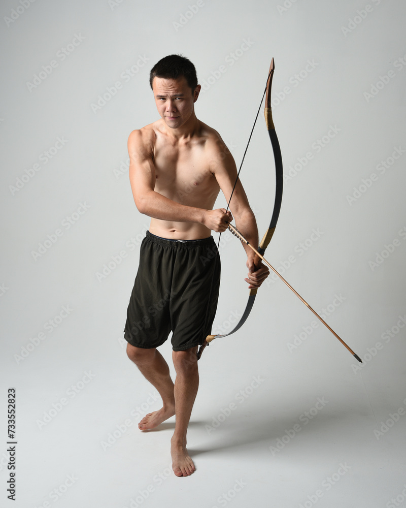 Full length portrait of fit asian male model, Holding hunting bow and arrow archery weapon, standing in warrior training action pose, isolated on white studio background. Full length portrait of fit asian male model, Holding hunting bow and arrow archery weapon, standing in warrior training action pose, isolated on white studio background.