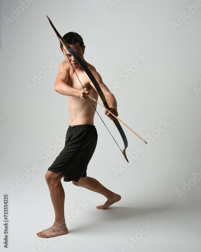 Full length portrait of fit asian male model, Holding hunting bow and arrow archery weapon, standing in warrior training action pose, isolated on white studio background.