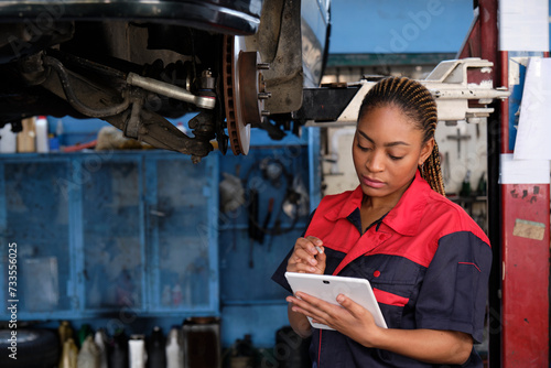 Expert young Black female automotive mechanic worker checks car's wheel brake disc and repairs inspect checklist by tablet at fix garage. Vehicle maintenance service works industry occupation jobs.