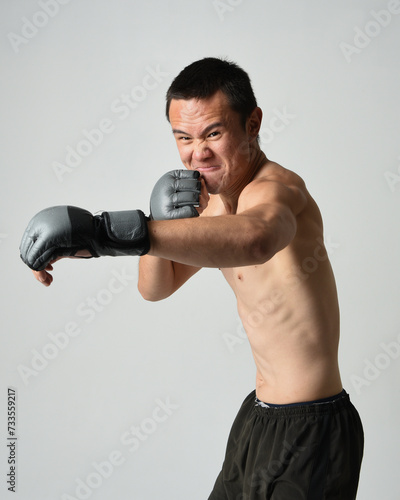 Close up portrait of fit asian male model, shirtless with muscles. Wearing gym shorthand boxing gloves, gestural punching pose. Isolated on a white studio background.