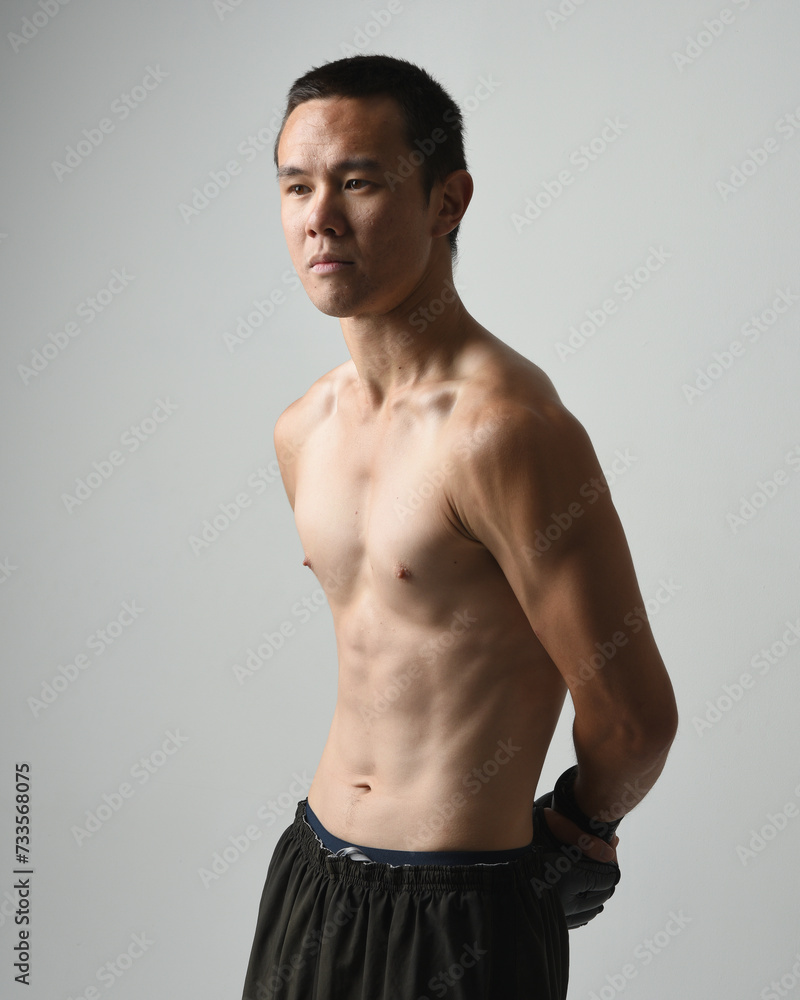 Fototapeta premium Close up portrait of fit asian male model, shirtless with muscles. Wearing gym shorthand boxing gloves, gestural punching pose. Isolated on a white studio background.