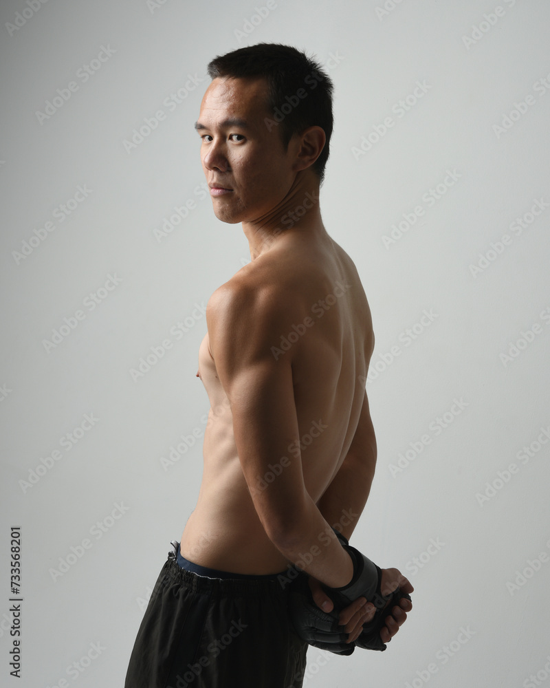 Fototapeta premium Close up portrait of fit asian male model, shirtless with muscles. Wearing gym shorthand boxing gloves, gestural punching pose. Isolated on a white studio background.