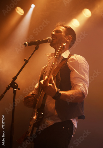 Concert, singer and guitar with man on stage for music, performance and rock show. Event, spotlight and energy with male musician playing instrument at festival club for rave, disco and celebration