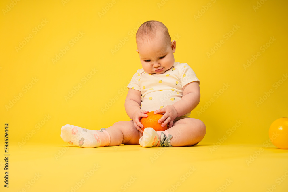 Beautiful baby girl holds orange sitting in studio on yellow background. Beautiful baby girl holds orange sitting in studio on yellow background.