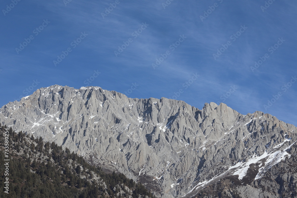 Rugged mountain range with sharp, rocky peaks under a clear blue sky. Patches of snow cling to the crevices of the mountainside, and a sparse forest is visible at the lower elevations.