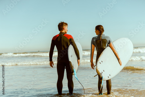 Back view of couple of surfers with surfboards standing on the beach and looking each other