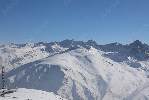 vast expanse of snow-covered mountains under a bright blue sky. The terrain is undulating with smooth snowfields and rugged peaks, creating a serene and cold landscape. © Mohammed Ajzal