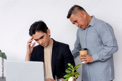 A stern and overbearing boss puts pressure on a visibly stressed man to complete deadlines on his work. A tough and taxing work environment. Negative effects of micromanagement. photo