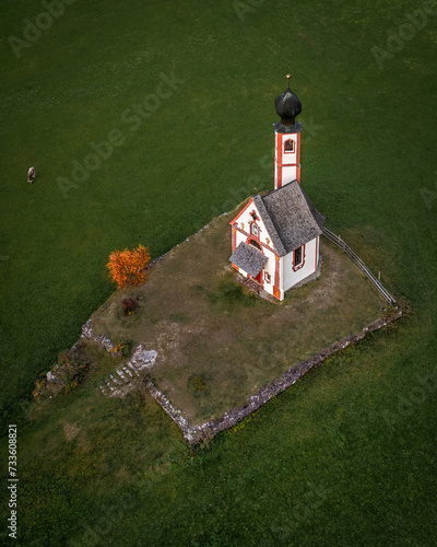 Val Di Funes, Dolomites, Italy - Aerial view of the beautiful St. Johann Church (Chiesetta di San Giovanni in Ranui) at South Tyrol with green grass, cow and autumn foliage photo