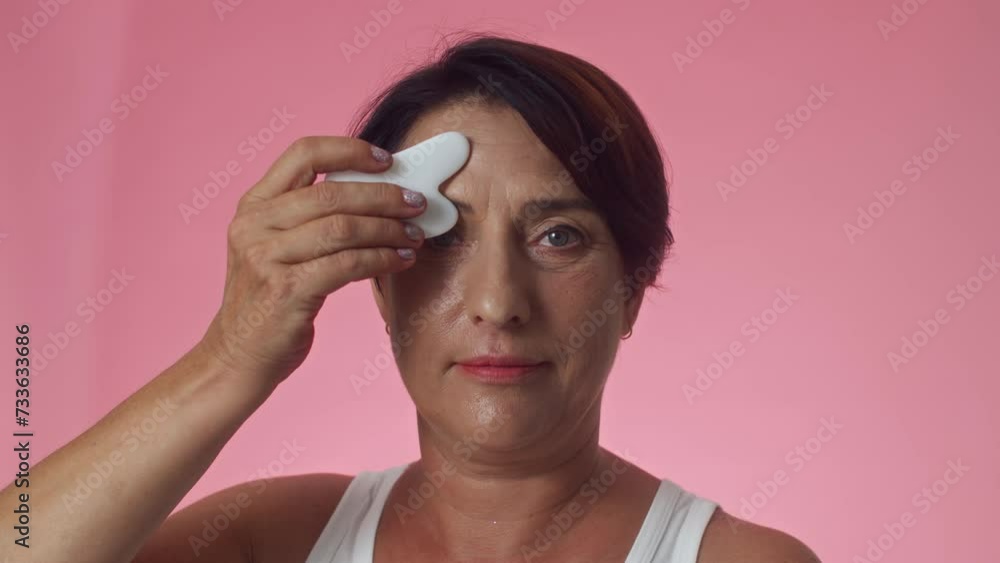 Medium close up of mature Caucasian woman in white top doing facial massage with gua sha tool and looking at camera Medium close up of mature Caucasian woman in white top doing facial massage with gua sha tool and looking at camera