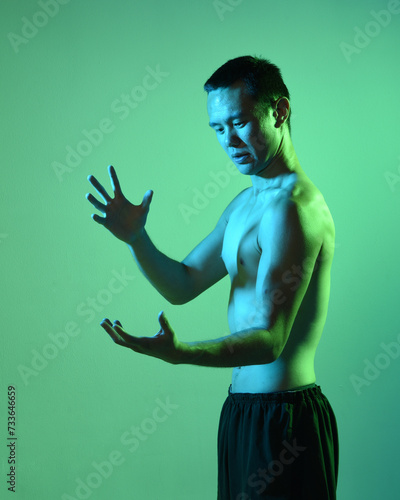 Fototapeta Naklejka Na Ścianę i Meble -  Close up portrait of fit asian male model, shirtless with muscles.  gestural ti chi inspired posing with arms reaching out  Isolated on a moody dark green studio background with silhouette.