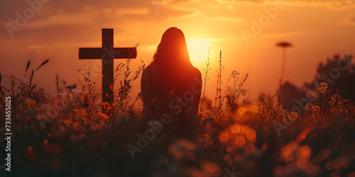 Silhouette of a woman sitting on the grass praying in front of a cross at sunset