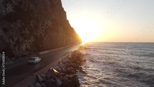 This cinematic footage is made with a drone, showing the car passing the road which is located near the sea and under a cliff, all this is happening during the sunset.