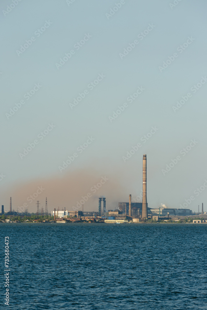 Industrial skyline dominated by a metallurgical plants tall chimneys emitting smoke over a body of water, symbolizing industrial pollution and eco awareness issues.