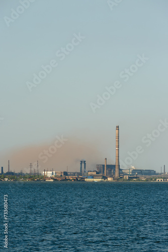 Industrial skyline dominated by a metallurgical plants tall chimneys emitting smoke over a body of water, symbolizing industrial pollution and eco awareness issues.