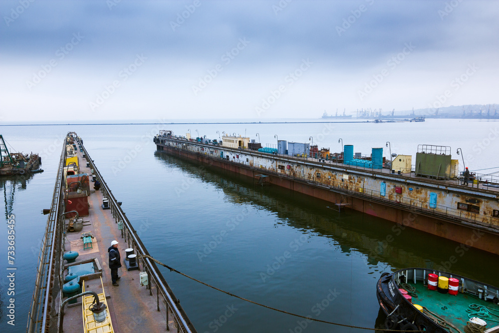Vessel maneuvers into floating dock at maritime yard for repairs, cloudy skies, surrounded by port infrastructure amidst calm waters. Specialist crew prepare for docking, maritime operations.