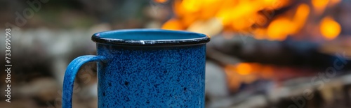 Header of a blue metal enamel mug stands on the stone with campfire in the mountains on background