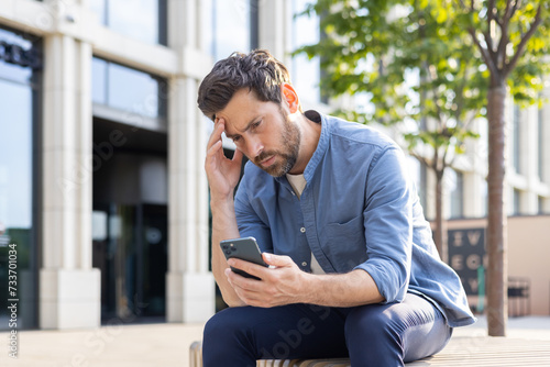 An upset young man sits on a bench on a city street and looks disappointedly at the screen of the phone he is holding in his hands