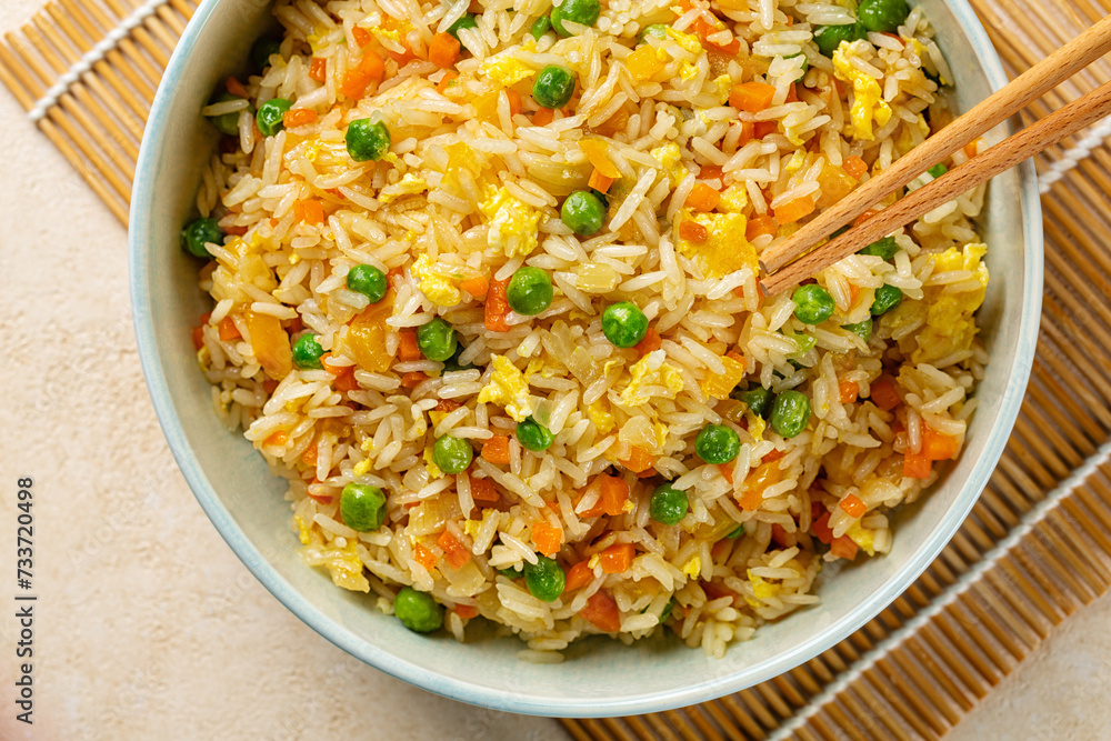 Top view of a bowl with fried rice with egg and vegetables, onion, bell pepper, carrot, green peas. Prepared and served in a light blue plate on a beige table. Top view of a bowl with fried rice with egg and vegetables, onion, bell pepper, carrot, green peas. Prepared and served in a light blue plate on a beige table.