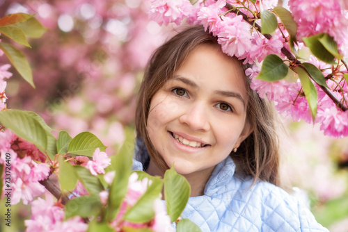 Young smile girl in a trench coat smiles and dreams standing at blossoming sakura garden. Outdoor fashion portrait little girl wearing stylish blue trench coat in blossom spring park. 