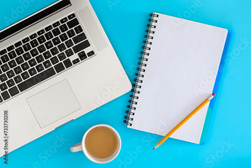 Blank notebook, laptop and cup of coffee on a blue background, top view.