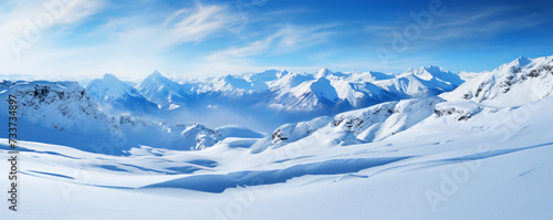 Panoramic view of a snowy mountain range. The mountains are covered in snow and the valley is surrounded by trees. The sky is a clear blue and there are a few clouds in the distance. © Rabbit_1990