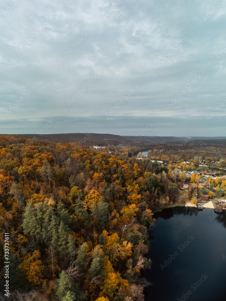 Autumn aerial vertical panorama of river valley with grey clouds. Flying above autumnal vibrant trees near rural riverside in Ukraine