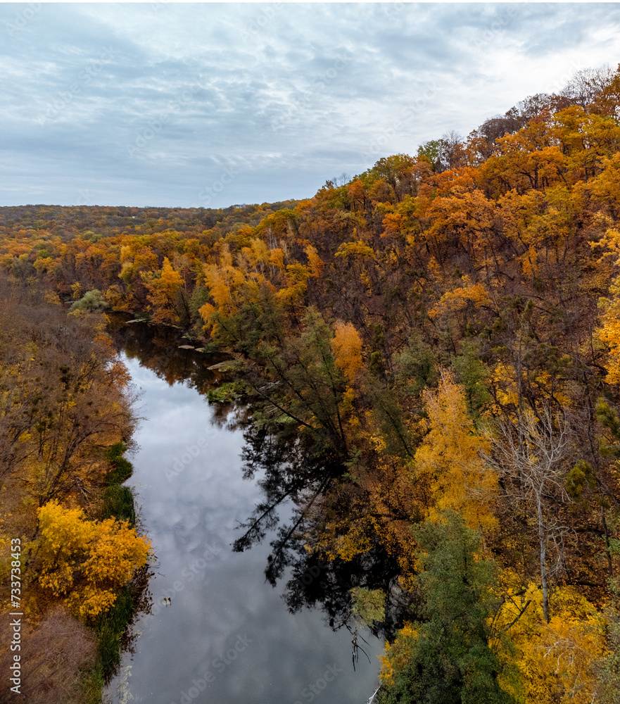 Autumn aerial river and colorful golden trees forest with grey cloudy sky. Flying above autumnal riverside in Ukraine